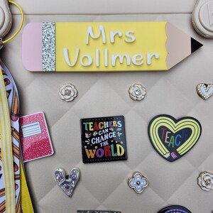 K&ouml;nnte beinhalten: Eine Sammlung von Accessoires zum Thema Lehrer, darunter ein gelbes, bleistiftf&ouml;rmiges Namensschild mit "Mrs Vollmer" in Wei&szlig; und verschiedene dekorative Charms. Zu den Charms geh&ouml;ren ein Notizbuch, ein Herz und ein Quadrat mit dem Text "Teachers Can Change the World".