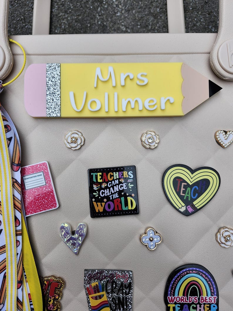 K&ouml;nnte beinhalten: Eine Sammlung von Accessoires zum Thema Lehrer, darunter ein gelbes, bleistiftf&ouml;rmiges Namensschild mit "Mrs Vollmer" in Wei&szlig; und verschiedene dekorative Charms. Zu den Charms geh&ouml;ren ein Notizbuch, ein Herz und ein Quadrat mit dem Text "Teachers Can Change the World".