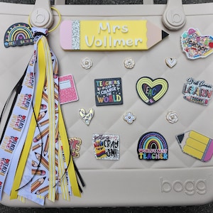 K&ouml;nnte beinhalten: Eine beige Tragetasche, verziert mit Accessoires zum Thema Lehrer. Die Tasche hat ein bleistiftf&ouml;rmiges Namensschild mit der Aufschrift "Mrs. Vollmer" sowie verschiedene Anstecknadeln und Anh&auml;nger. Eine Quaste mit Bleistift- und "Teach Love Inspire"-B&auml;ndern h&auml;ngt am Griff. Die Tasche ist eine Bogg Bag.
