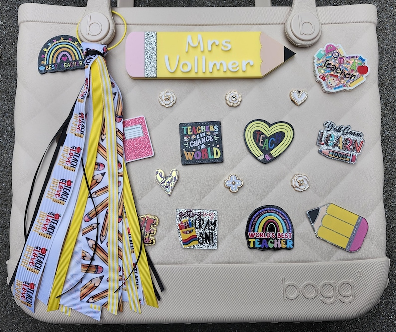 K&ouml;nnte beinhalten: Eine beige Tragetasche, verziert mit Accessoires zum Thema Lehrer. Die Tasche hat ein bleistiftf&ouml;rmiges Namensschild mit der Aufschrift "Mrs. Vollmer" sowie verschiedene Anstecknadeln und Anh&auml;nger. Eine Quaste mit Bleistift- und "Teach Love Inspire"-B&auml;ndern h&auml;ngt am Griff. Die Tasche ist eine Bogg Bag.