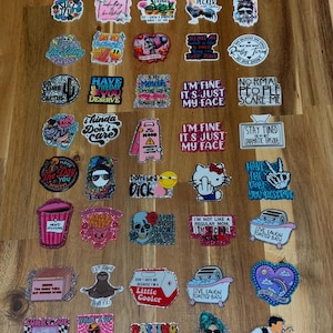 May include: A collection of colorful stickers with various sayings and designs. The stickers feature glitter and are arranged in rows on a wooden surface. Some of the sayings include "I'm fine it's just my face", "Have the day you deserve", "Don't be a dick", and "Live laugh toasted bath".