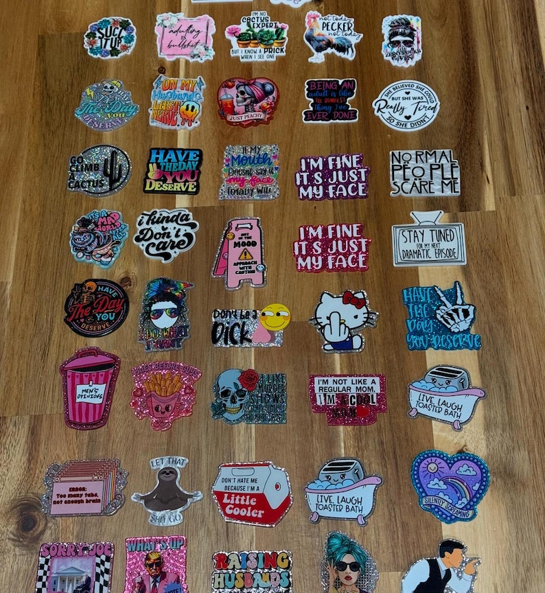 May include: A collection of colorful stickers with various sayings and designs. The stickers feature glitter and are arranged in rows on a wooden surface. Some of the sayings include "I'm fine it's just my face", "Have the day you deserve", "Don't be a dick", and "Live laugh toasted bath".
