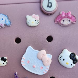 K&ouml;nnte beinhalten: Eine Sammlung farbenfroher dekorativer Charms mit Zeichentrickfiguren, darunter Hello Kitty und My Melody, die an einer rosa Tasche befestigt sind. Die Charms sind in verschiedenen Farben und Designs gehalten und verleihen einen verspielten Touch.