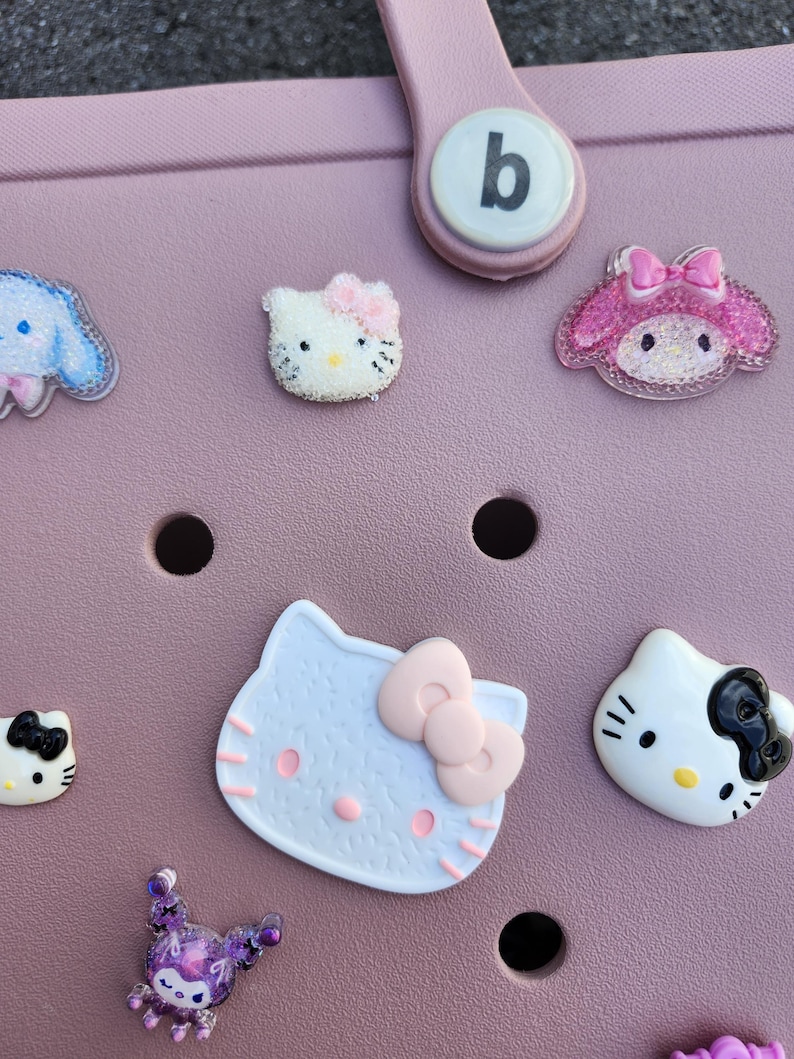 K&ouml;nnte beinhalten: Eine Sammlung farbenfroher dekorativer Charms mit Zeichentrickfiguren, darunter Hello Kitty und My Melody, die an einer rosa Tasche befestigt sind. Die Charms sind in verschiedenen Farben und Designs gehalten und verleihen einen verspielten Touch.
