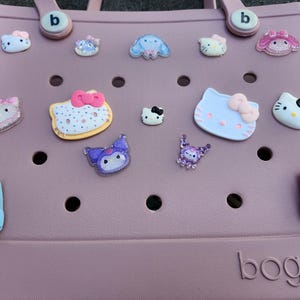 K&ouml;nnte beinhalten: Eine hellrosa Tragetasche mit verschiedenen dekorativen Anh&auml;ngern. Die Anh&auml;nger zeigen Zeichentrickfiguren und Designs in Farben wie Rosa, Blau und Lila. Die Tasche hat einen Griff und der Markenname "bogg" ist sichtbar.