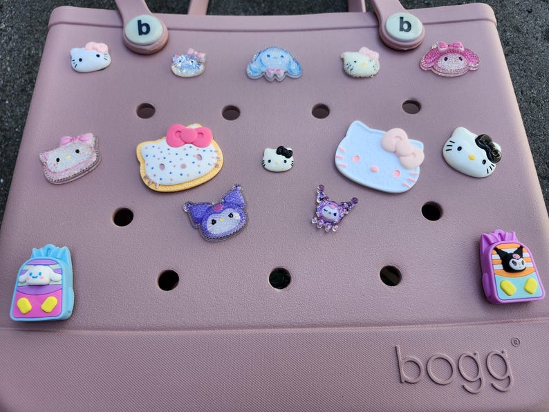 K&ouml;nnte beinhalten: Eine hellrosa Tragetasche mit verschiedenen dekorativen Anh&auml;ngern. Die Anh&auml;nger zeigen Zeichentrickfiguren und Designs in Farben wie Rosa, Blau und Lila. Die Tasche hat einen Griff und der Markenname "bogg" ist sichtbar.