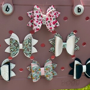 May include: Five different hair bows displayed on a pink surface. The bows are made of fabric and have various patterns, including floral, polka dots, and owls. The bows are all different sizes and shapes.