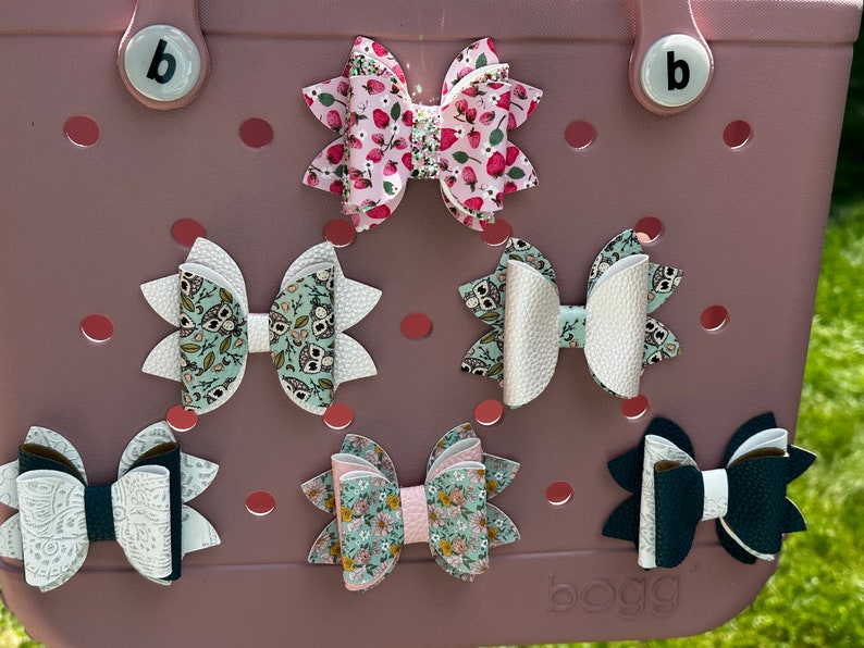 May include: Five different hair bows displayed on a pink surface. The bows are made of fabric and have various patterns, including floral, polka dots, and owls. The bows are all different sizes and shapes.