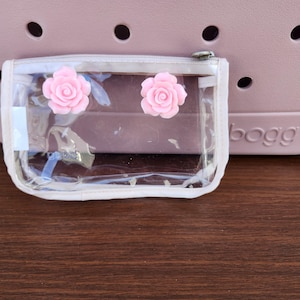 May include: A clear, zippered pouch with two pink rose embellishments. The pouch is set against a light pink tote bag with the word "bogg" visible. The background is a wooden surface.