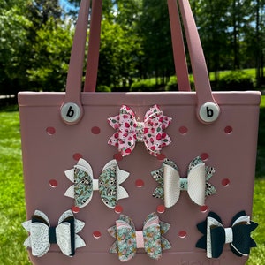 May include: A pink and white tote bag with the word "boga" printed on the bottom. The bag has six different patterned hair bows attached to it.