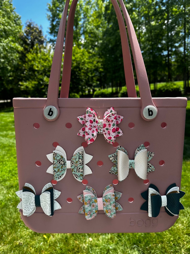 May include: A pink and white tote bag with the word "boga" printed on the bottom. The bag has six different patterned hair bows attached to it.