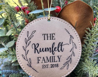 May include: A round, tan leather ornament with the text "THE Rumfelt FAMILY EST. 2018" in black script and a decorative leaf design. The ornament hangs from a gold string and is surrounded by greenery and red berries.