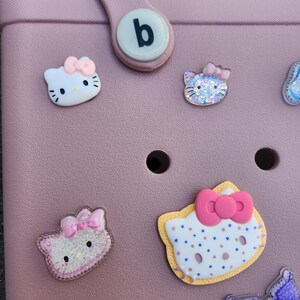 K&ouml;nnte beinhalten: Eine rosa Tragetasche mit mehreren dekorativen Hello Kitty-Anh&auml;ngern. Die Anh&auml;nger haben verschiedene Gr&ouml;&szlig;en und Designs, mit Schleifen und Glitzerakzenten. Ein wei&szlig;er runder Knopf mit einem schwarzen "b" befindet sich am Griff.