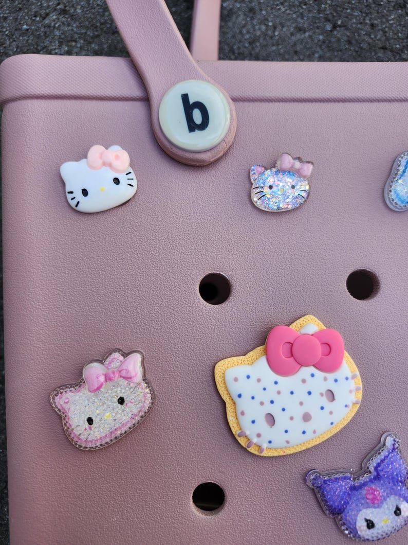 K&ouml;nnte beinhalten: Eine rosa Tragetasche mit mehreren dekorativen Hello Kitty-Anh&auml;ngern. Die Anh&auml;nger haben verschiedene Gr&ouml;&szlig;en und Designs, mit Schleifen und Glitzerakzenten. Ein wei&szlig;er runder Knopf mit einem schwarzen "b" befindet sich am Griff.