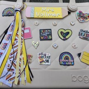 K&ouml;nnte beinhalten: Beige Tragetasche mit Lehrer-Themen-Anh&auml;ngern und einem Quastenanh&auml;nger. Zu den Anh&auml;ngern geh&ouml;ren ein Bleistift, ein Notizbuch, ein Herz und ein Regenbogen. Die Tasche hat ein Steppmuster und das Wort "bogg" auf der Unterseite.
