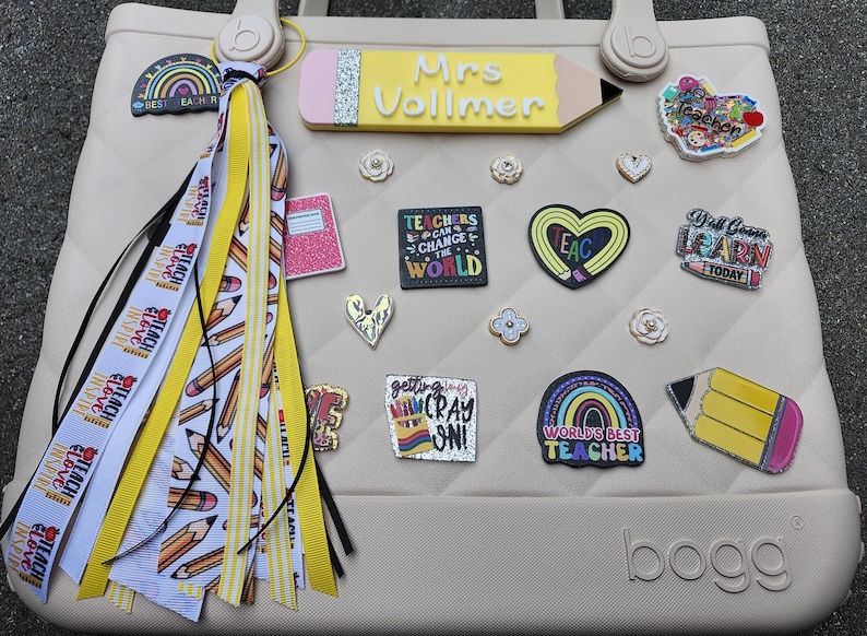 K&ouml;nnte beinhalten: Beige Tragetasche mit Lehrer-Themen-Anh&auml;ngern und einem Quastenanh&auml;nger. Zu den Anh&auml;ngern geh&ouml;ren ein Bleistift, ein Notizbuch, ein Herz und ein Regenbogen. Die Tasche hat ein Steppmuster und das Wort "bogg" auf der Unterseite.