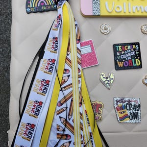 K&ouml;nnte beinhalten: Eine beige Tasche mit Accessoires zum Thema Lehrer. Eine Quaste mit B&auml;ndern mit der Aufschrift "Teach, Love, Inspire" und Bleistiftgrafiken h&auml;ngt an der Tasche. Weitere Accessoires sind ein namensschildf&ouml;rmiger Bleistift, ein Regenbogen-Charm und Anstecknadeln mit inspirierenden Zitaten.