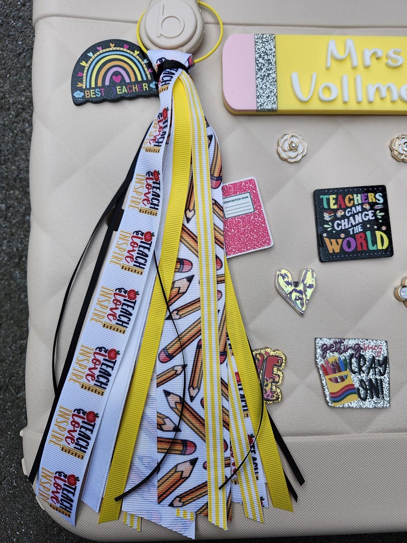 K&ouml;nnte beinhalten: Eine beige Tasche mit Accessoires zum Thema Lehrer. Eine Quaste mit B&auml;ndern mit der Aufschrift "Teach, Love, Inspire" und Bleistiftgrafiken h&auml;ngt an der Tasche. Weitere Accessoires sind ein namensschildf&ouml;rmiger Bleistift, ein Regenbogen-Charm und Anstecknadeln mit inspirierenden Zitaten.