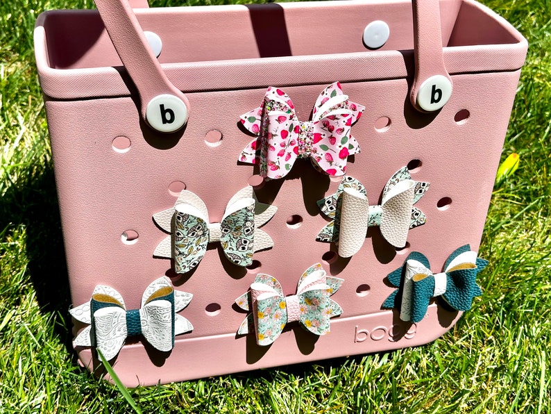May include: A pink tote bag with a white handle and a white letter "b" on the side. The bag has a grid of holes and is decorated with six colorful fabric bows.