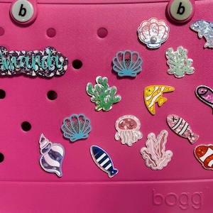 May include: A pink plastic tote bag with the word "BOGG" printed on the bottom. The bag is decorated with various colorful stickers, including seashells, fish, seaweed, and a jellyfish. The name "Amanda" is written in glitter on the top of the bag.
