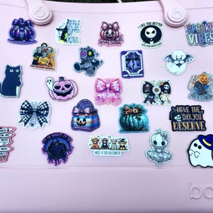 May include: A collection of Halloween-themed charms and stickers on a pink tote bag. The charms feature various designs, including ghosts, pumpkins, bows, and text like "Spooky Vibes" and "Have the day you deserve".