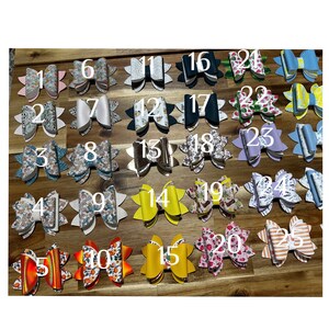 May include: Twenty-five different hair bows with various patterns and colors. The bows are arranged in five rows of five. The bows are made of fabric and have a variety of designs, including floral, geometric, and abstract patterns.