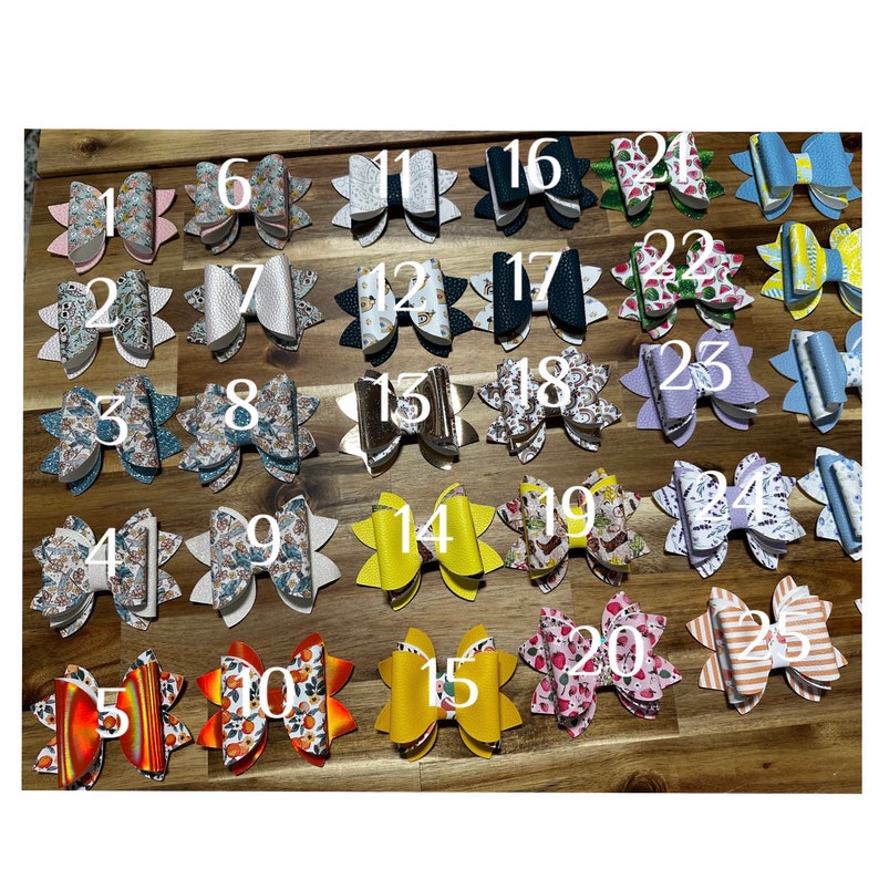 May include: Twenty-five different hair bows with various patterns and colors. The bows are arranged in five rows of five. The bows are made of fabric and have a variety of designs, including floral, geometric, and abstract patterns.
