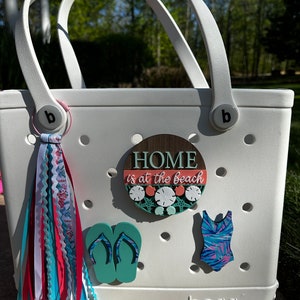 May include: White tote bag with the word "BOGG" printed on the bottom. The bag has a variety of colorful accessories attached to it, including a wooden round sign that says "HOME is at the beach", a pair of flip flops, a swimsuit, and a long, colorful ribbon.