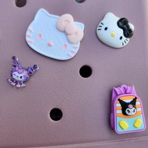 K&ouml;nnte beinhalten: Eine Sammlung bunter Schuhanh&auml;nger mit Zeichentrickfiguren. Enth&auml;lt einen hellblauen Hello Kitty-Kopf mit rosa Schleife, einen wei&szlig;en Hello Kitty-Kopf mit schwarzer Schleife, ein lila Wesen und einen Rucksack mit einer Zeichentrickfigur.