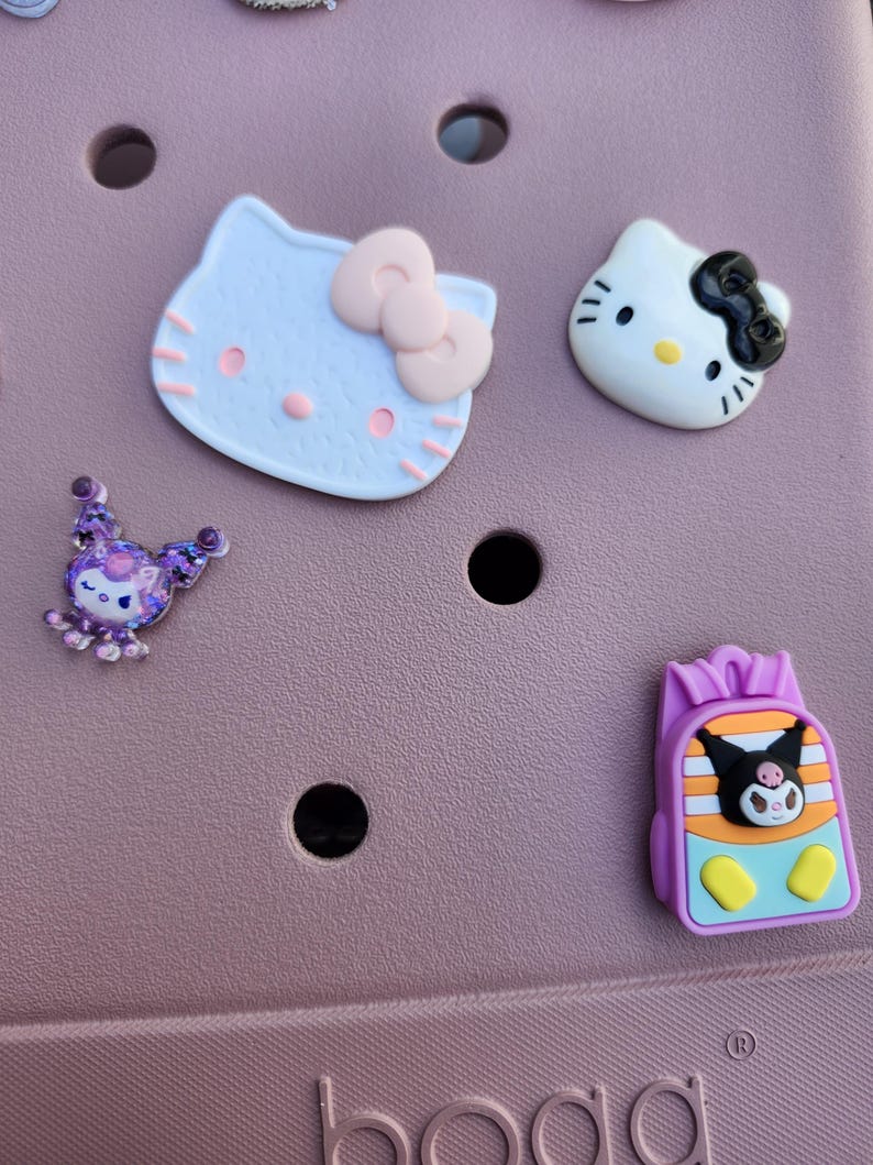K&ouml;nnte beinhalten: Eine Sammlung bunter Schuhanh&auml;nger mit Zeichentrickfiguren. Enth&auml;lt einen hellblauen Hello Kitty-Kopf mit rosa Schleife, einen wei&szlig;en Hello Kitty-Kopf mit schwarzer Schleife, ein lila Wesen und einen Rucksack mit einer Zeichentrickfigur.