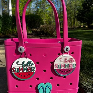 May include: A bright pink tote bag with two decorative charms that say "Hello Summer" and a pair of turquoise flip-flop charms. The charms feature watermelon designs. The bag is sitting on a stone surface.