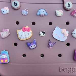 K&ouml;nnte beinhalten: Eine staubrosa Tragetasche mit verschiedenen dekorativen Anh&auml;ngern. Die Anh&auml;nger zeigen Zeichentrickfiguren, darunter Hello Kitty, in verschiedenen Farben. Auf der Tasche steht das Wort "bogg" auf der Unterseite.