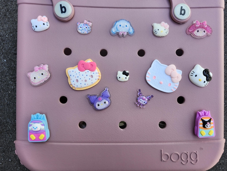 K&ouml;nnte beinhalten: Eine staubrosa Tragetasche mit verschiedenen dekorativen Anh&auml;ngern. Die Anh&auml;nger zeigen Zeichentrickfiguren, darunter Hello Kitty, in verschiedenen Farben. Auf der Tasche steht das Wort "bogg" auf der Unterseite.