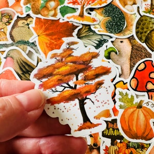 Op de afbeelding: Een sticker met een aquarel-illustratie van een boom met oranje, gele en bruine bladeren. De boom is omringd door kleine witte stippen die lijken op vallende bladeren. De sticker is op een witte achtergrond.