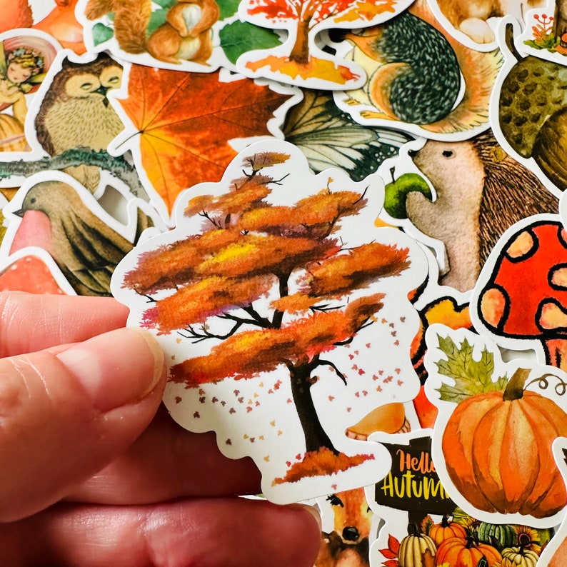 Op de afbeelding: Een sticker met een aquarel-illustratie van een boom met oranje, gele en bruine bladeren. De boom is omringd door kleine witte stippen die lijken op vallende bladeren. De sticker is op een witte achtergrond.