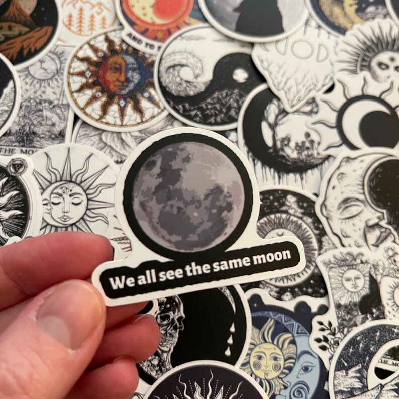 May include: A black and white sticker with a gray moon and the text "We all see the same moon". The sticker is surrounded by other stickers with various celestial designs.