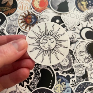 May include: A white sticker with a black and white illustration of a sun with a crescent moon inside. The sun has a face with closed eyes and the moon has a face with a single eye.