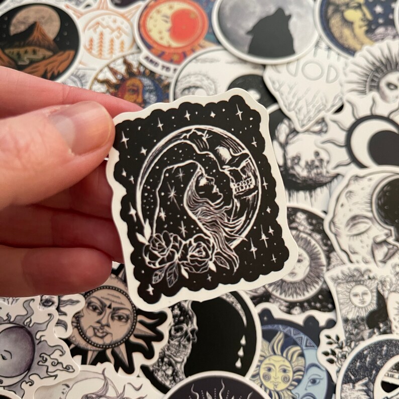 May include: A black and white sticker with a moon and a woman's face. The sticker is surrounded by stars and has a floral design at the bottom.