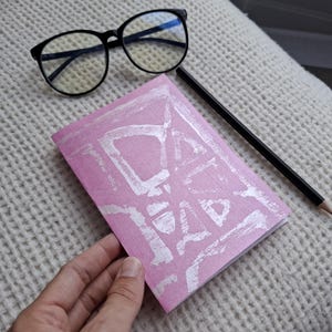 May include: A pink notebook with a white block-printed design on the cover, accompanied by a pair of black-rimmed spectacles and a black pencil. The notebook features a textured surface and a rectangular shape with a hand-printed graphic pattern.