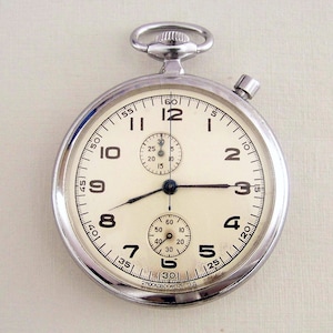 Puede incluir: Un reloj de bolsillo plateado con una esfera blanca y agujas negras. El reloj tiene una función de cronógrafo y un pequeño subdial para los segundos. El reloj está marcado con "Московский ЧАС З Д".