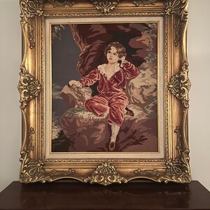 May include: A framed needlepoint tapestry depicting a young person in a red outfit sitting on a rock. The tapestry is framed in a gold ornate frame.