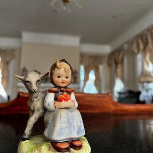 May include: A porcelain figurine of a girl in a blue and white dress holding a bouquet of red flowers. She is sitting next to a small, gray goat.