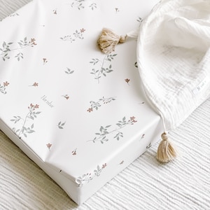 May include: White changing pad cover with a floral pattern featuring small red flowers and grey leaves. The cover has a white tassel on each corner and the word "Morishor" printed in the centre.