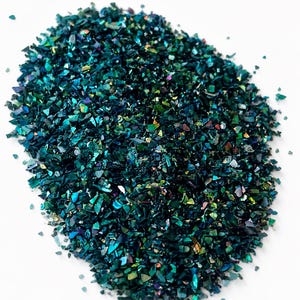 Könnte beinhalten: Eine Ansammlung kleiner, unregelmäßiger, zerkleinerter Muschelfragmente in tiefen Blau-, Smaragdgrün- und Petroltönen. Die Stücke haben eine reflektierende, schillernde Oberfläche, die das Licht einfängt und einen schimmernden Effekt erzeugt.
