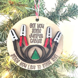 May include: A wooden Christmas ornament with the text "GOT YOU SOME JUMPER CABLES Since you like to start shit." The ornament features a design of jumper cables with red and black details, and a small green Christmas tree.