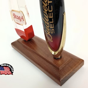 May include: Two beer tap handles on a wooden base. The left handle is clear with a red base and says "Stag" in gold lettering. The right handle is black with a red and gold base and says "Budweiser Select" in gold lettering. The base is made in the USA.