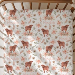 May include: A white fitted crib sheet with a repeating pattern of brown Highland cows, pink flowers, and green leaves. The name "Marabelle" is written in a cursive font throughout the design. The sheet is displayed in a wooden crib.