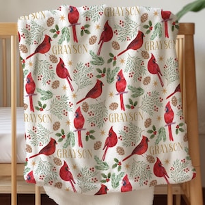 May include: A cream-colored baby blanket with a repeating pattern of red cardinals, pinecones, and greenery. The name "Grayson" is printed throughout the design. The blanket is draped over a wooden crib.