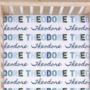 May include: A white crib sheet with the name "Theodore" printed repeatedly in blue and green. The crib sheet is inside a light wood crib.
