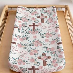 May include: A white changing pad with a floral pattern of pink roses and green leaves. Brown crosses are interspersed throughout the design. The name "Charlotte Rose" is written in cursive script on the pad.